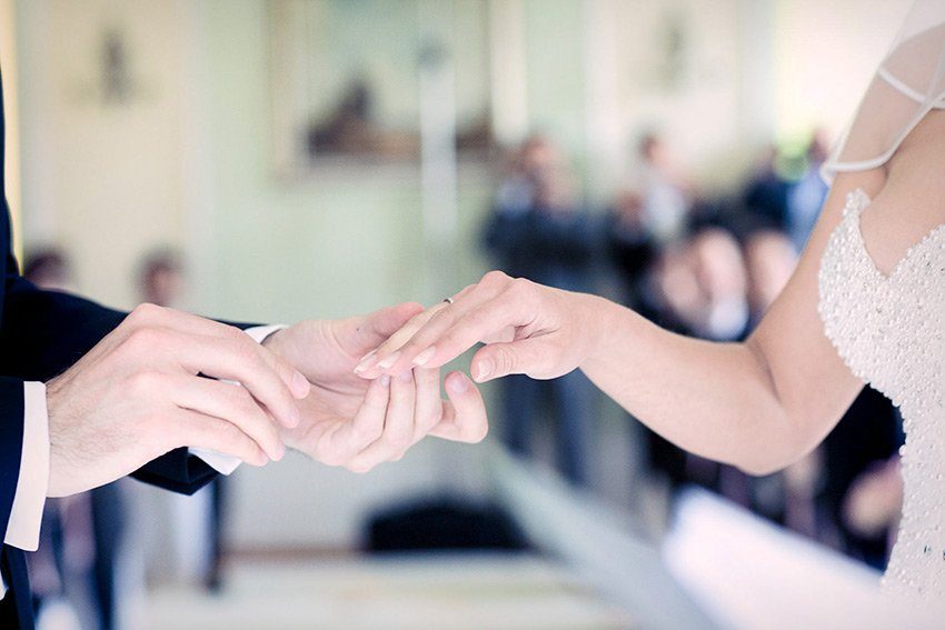 Nahaufnahme eines Brautpaars, das sich während der Trauung die Ringe ansteckt, mit unscharfen Gästen im Hintergrund.

