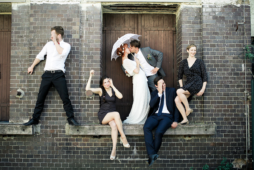 Brautpaar, das sich vor einer Backsteinmauer küsst, umgeben von spielerisch posierender Hochzeitsgesellschaft.