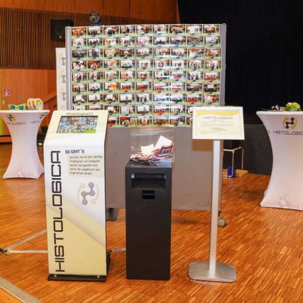 Ein Sofortdruck-Fotostand mit einem Drucker und einer Pinnwand voller Fotos auf einer Veranstaltung.