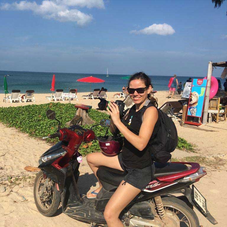 Hochzeitsfotografin auf einem Motorroller am Strand von Koh Lanta, Thailand, mit Liegestühlen und Sonnenschirmen im Hintergrund