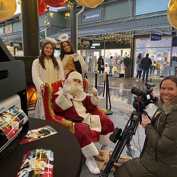 Mobiler Fotograf mit Fotodruck vor Ort im Einkaufscenter, Weihnachtsmann in rotem Sessel, umgeben von zwei Engeln und einer Fotografin.