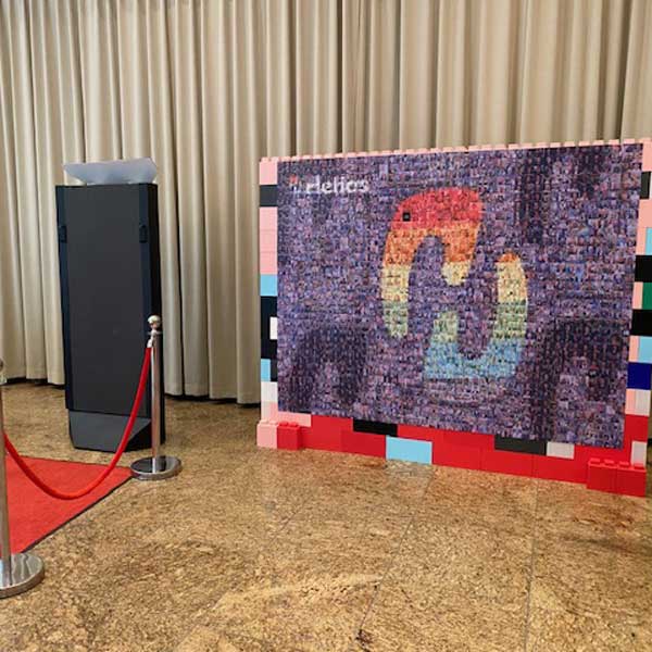 Fotomosaic Wall als interaktives Fotoerlebnis auf einem Event, eingerahmt von bunten Bausteinen und begleitet von einem Fotospiegel mit rotem Teppich.
